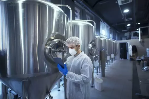 Técnico com touca, máscara e tablet ao lado de tanques de aço inox em fábrica de bebidas