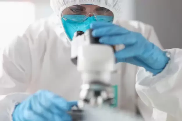 Técnica com roupa de proteção, máscara e luvas ajustando um microscópio em laboratório