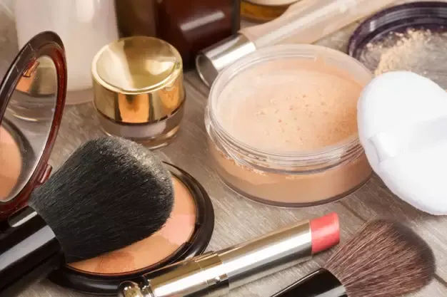 Produtos cosméticos sobre bancada: pó facial, pincéis, batom e frascos