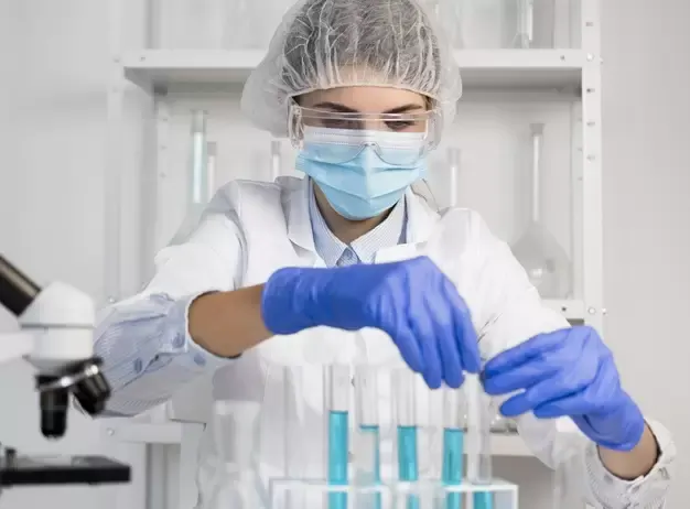 Técnica com touca, óculos, máscara e luvas manuseando tubos de ensaio em laboratório