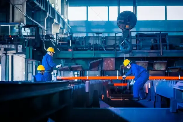 Operadores com capacete em fábrica metalúrgica com ponte rolante e barras metálicas aquecidas