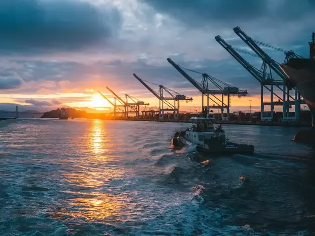 Rebocador navegando em porto com guindastes ao pôr do sol